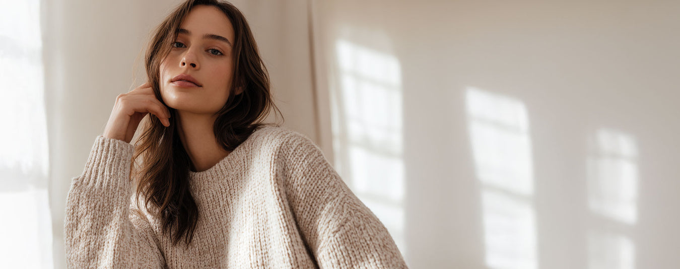 Woman wearing a beige sweater standing in a softly lit room with sunlight filtering through.